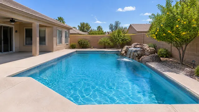 Backyard of a single-story early-2000s Gilbert, Arizona stucco home: rectangular pool with a fresh blue plaster finish, a short rock waterfall, cool-deck patio and a lemon tree against a tan block wall