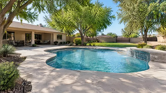 Large mature backyard of a single-story ranch-style Gilbert, Arizona home: older free-form pool being refreshed with new tile and a wide deck, big shade trees, RV gate and a grassy yard beyond