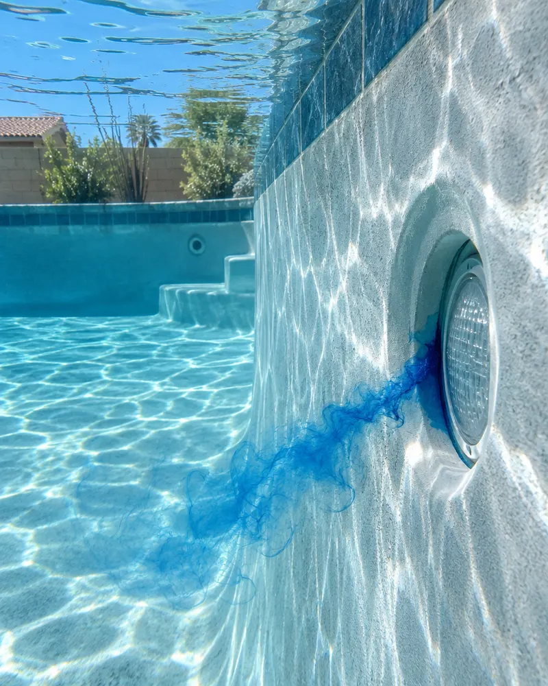Underwater view of a pool wall with a fine dye stream drifting into a gap around a light niche, bright plaster finish, sun rays