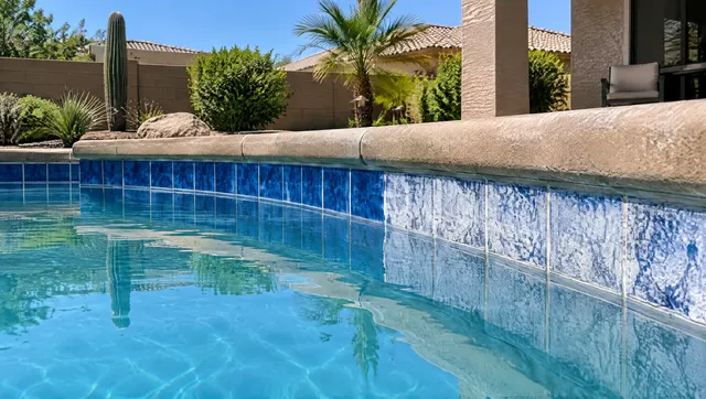 Sparkling clean blue and white ceramic waterline tile on a backyard pool after calcium removal, half the band still showing white scale for contrast, calm water, tan coping, Gilbert, Arizona