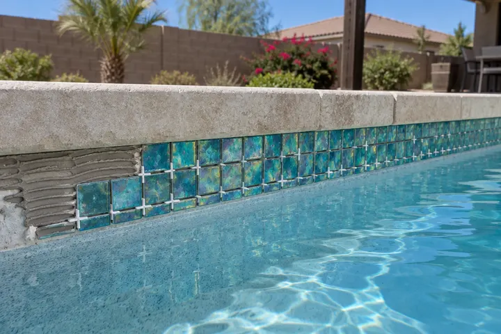New iridescent teal glass mosaic tile being set along a pool waterline, fresh thinset and spacers, coping above